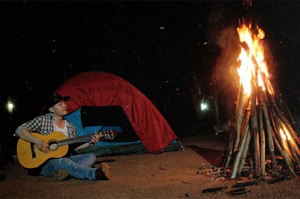 Chàng trai ôm đàn guitar ngồi cạnh lều và lửa trại